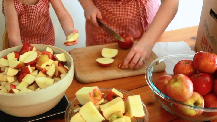 woman chopping apples on wood cutting board with toddler girl adding them to bowl